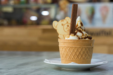 White plate with ice cream dessert in wafer cup with chocolate cookies and creative decoration topping on blurred colorful interior background.