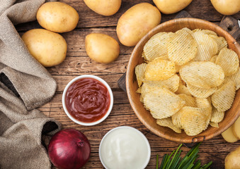 Fresh organic homemade potato crisps chips in wooden bowl with sour cream and red onions and spices on wooden table background. Top viewFresh yellow potatoes with ketchup