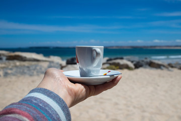 a cup of coffee in wpmans hand with view on beautiful beach and blue sky