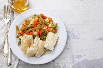 boiled cod fish with boiled vegetables on white plate on ceramic background