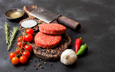 Fresh raw minced homemade farmers grill beef burgers on vintage chopping board with spices and herbs and meat hatchet on black board. Oil, garlic and tomatoes.