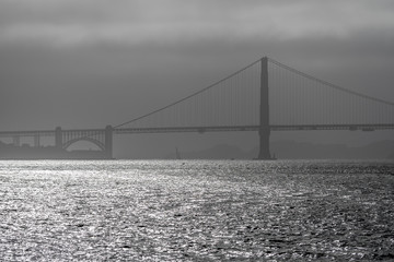 Monochrome view of San Francisco Bay looking west to the Golden Gate Bridge on a partly sunny late afternoon.  Bright water, dark grey fog behind the bridge tower.