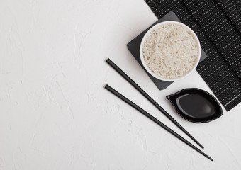 Fototapeta na wymiar White bowl with boiled organic basmati jasmine rice with black chopsticks and sweet soy sauce on bamboo place mat on white background. Space for text