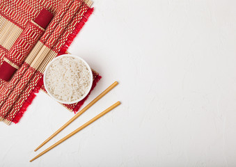 Fototapeta na wymiar White bowl with boiled organic basmati jasmine rice with wooden chopsticks and sweet soy sauce on bamboo placemat with red linen towel on black stone background.