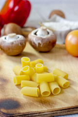 Dry rigatoni pasta on wooden kitchen board with vegetables.