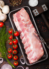 Plastic tray with raw pork ribs on chopping board and vintage meat fork and knife on wooden background. Fresh tomatoes and red onion with garlic, salt and pepper.