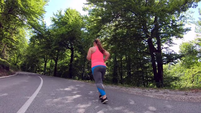 Wide low angle tracking shot of an overweight woman running on the sunny road in nature