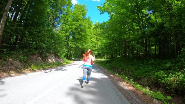 Wide forward tracking shot of an overweight woman running on the sunny road surrounded by trees in the summer