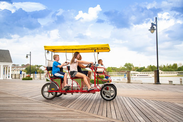 Young happy family riding a double surrey tandem bicycle on a large ocean boardwalk. Outdoor summer fun with kids - obrazy, fototapety, plakaty