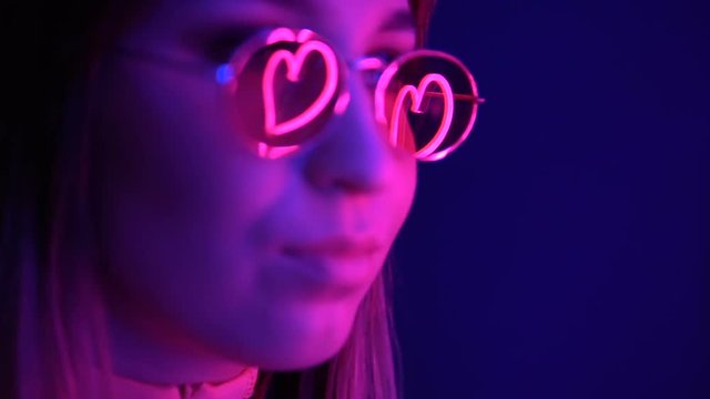 Fashionable girl in glasses on a neon background. Neon hearts are reflected in glasses.