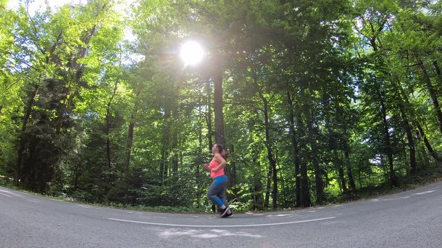 Locked down low angle shot of an overweight woman with pony tail running on an asphalt road through the forest in sunshine