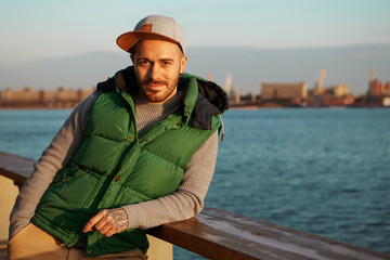 Obraz premium Style, urban fashion and people concept. Portrait of attractive confident young European guy with stubble, pierced eyebrow and tattoos on hands posing outdoors at wide river, having relaxed look