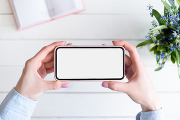 Female hands with a smartphone. White blank screen. Table with notebook and flowers on background