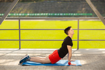 Young man training yoga outdoors. Sporty guy makes stretching exercise on a blue yoga mat, on the sports ground.