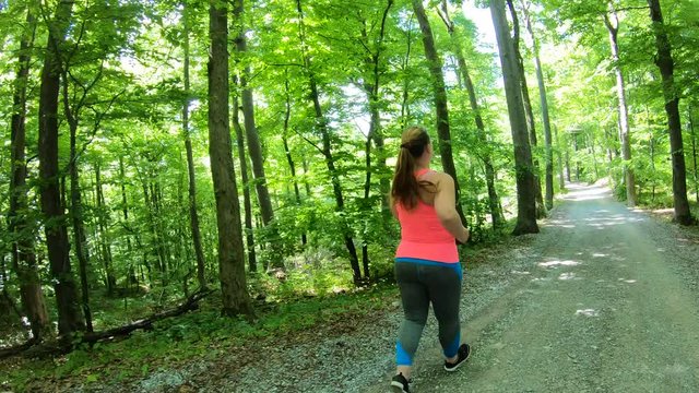 Forward tracking shot of an overweight woman with a pony tail running down the gravel forest road on a nice warm day