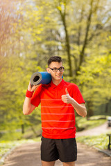 Young sports man in red t-shirt posing in a spring park with a blue yoga mat. His hands shows class