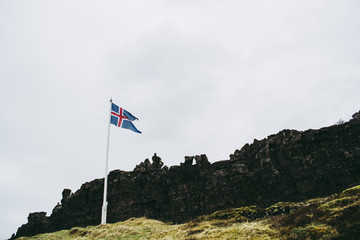 Flag of Iceland against natural background. Cold bleak landscapes of Thingvellir National Park in Iceland in spring. Ancient rocks against dark misty mountains and gray sky.