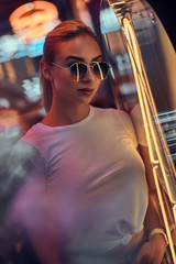 Portrait of pensive trendy woman in sunglasses and white tshirt with pink neon lights.