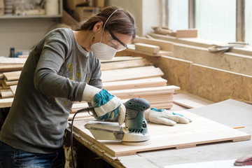 Young woman grinds the wood board with angular grinding machine at workshop
