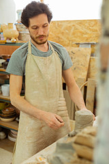 Focused dark-haired man in special apron measuring needed amount of clay