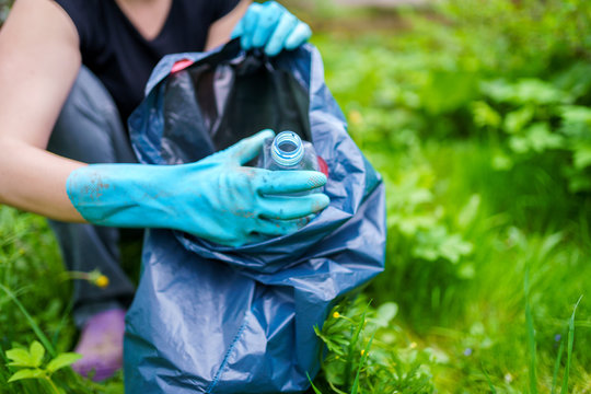 Image of woman in rubber gloves picking up dirty plastic bottle in bag on green lawn