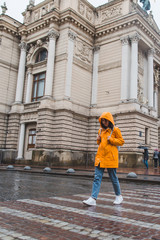Fototapeta na wymiar woman in orange raincoat crossing road old european building on background