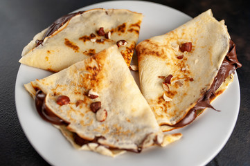 Pancakes with chocolate paste and hazelnuts, on a white plate on a background of concrete, slate