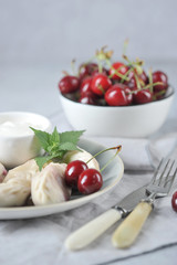 Dumplings with cherries and sour cream. A cup of cherries, cutlery and a napkin complement the composition. Light background. Vertical frame orientation.