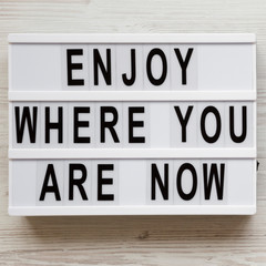 'Enjoy where you are now' words on a modern board over white wooden background, overhead view. Top view, flat lay, from above.