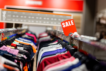 Retail clothes store clearance. Garment shop with various bright youth casual wear at discount price. Wear hangers and red SALE signboard. Seasonal sales on fashion market