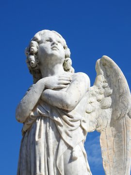 Scene in a graveyard: close-up of an old stone statue of an angel with arms crossed in the chest and looking at the sky. In the background, blue sky on a sunny day. Bottom view.
