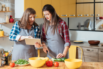 Online cooking class. Vegan healthy food recipe. Mother and daughter following culinary blog on tablet, preparing meal.