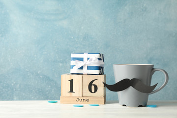 Obraz premium Cup with decorative mustache, gift box and wooden calendar on white table against blue background, space for text