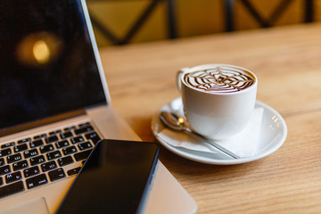 White Cup of cappuccino with pattern on the foam. Hot freshly brewed coffee in a white mug next to the smartphone and a laptop on a wooden table in the interior of the cafe.