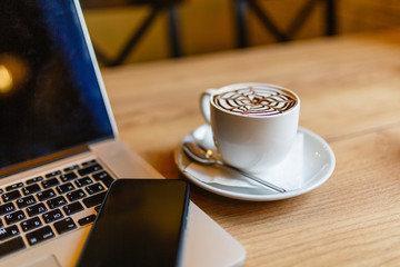 White Cup of cappuccino with pattern on the foam. Hot freshly brewed coffee in a white mug next to the smartphone and a laptop on a wooden table in the interior of the cafe.
