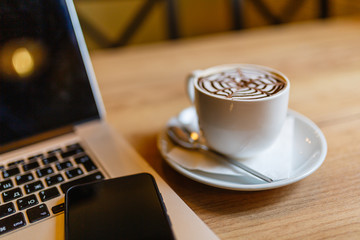 White Cup of cappuccino with pattern on the foam. Hot freshly brewed coffee in a white mug next to the smartphone and a laptop on a wooden table in the interior of the cafe.
