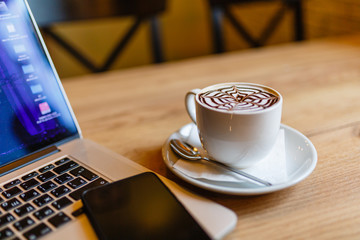 White Cup of cappuccino with pattern on the foam. Hot freshly brewed coffee in a white mug next to the smartphone and a laptop on a wooden table in the interior of the cafe.
