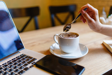 Women's hands holding a spoon over a Cup of coffee with pattern on the foam. Hot freshly brewed coffee in a white mug next to the smartphone and laptop on a wooden table in the interior of the cafe.