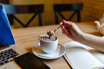 Women's hands holding spoon with foam over Cup of coffee with a pattern on the foam. Girl eats the foam from the cappuccino. Hot coffee in white mug next to the smartphone and laptop on wooden table.