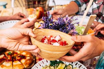 Point of view on wooden bowl with small tomatoes. Red checkered tablecloth in the table outdoor in the terrace. Wooden cutting board with pizza. Group of caucasian people enjoying meal together