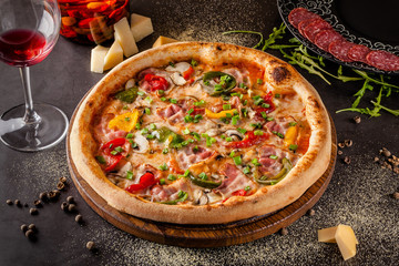 Concept Italian cuisine. A real pizza with vegetables and ham, bacon and parmesan cheese. The ingredients on the table. Red wine in a glass on the table. Closeup, background image