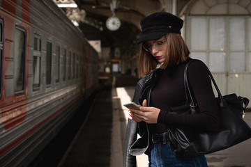 Attractive young woman millenial in black clothes and a hat at the railway station next to the train. Speaks on a mobile phone, smiles and waits for the train.