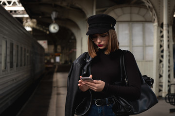 Attractive young woman millenial in black clothes and a hat at the railway station next to the train. Speaks on a mobile phone, smiles and waits for the train.