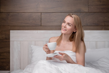 An attractive middle-aged blonde woman in a white bathrobe is lying on a bed in a bedroom. Drinking coffee from a white cup. Smiles and laughs.
