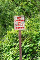 No camping or overnight parking sign with red lettering on rural road beside green trees.