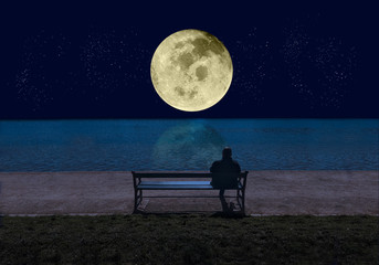 Man sitting on a bench by the sea in the  night and looking at a big shining full moon with stars...