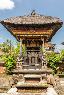 Dusun Ambengan, Bali, Indonesia - February 25, 2019: Decorated gray stone memorial pillar at clan compound in garden with green foliage under blue sky. Gold decorations, dark reed roof, statues of bea