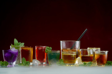 Color septet of alcoholic beverages in transparent glass glasses with ice and mint in smoke on a red background
