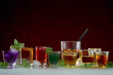 Color septet of alcoholic beverages in transparent glass glasses with ice and mint in smoke on a red background