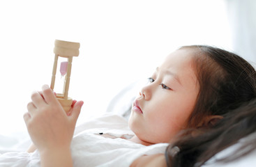 Portrait of little Asian girl looking at hourglass in hand lying on bed at home. Waiting times with sandglass. Close-up shot.